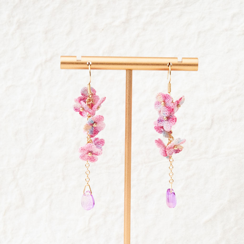 gold plated drop earrings with pink micro crochet flowers and a purple bead on a white textured background