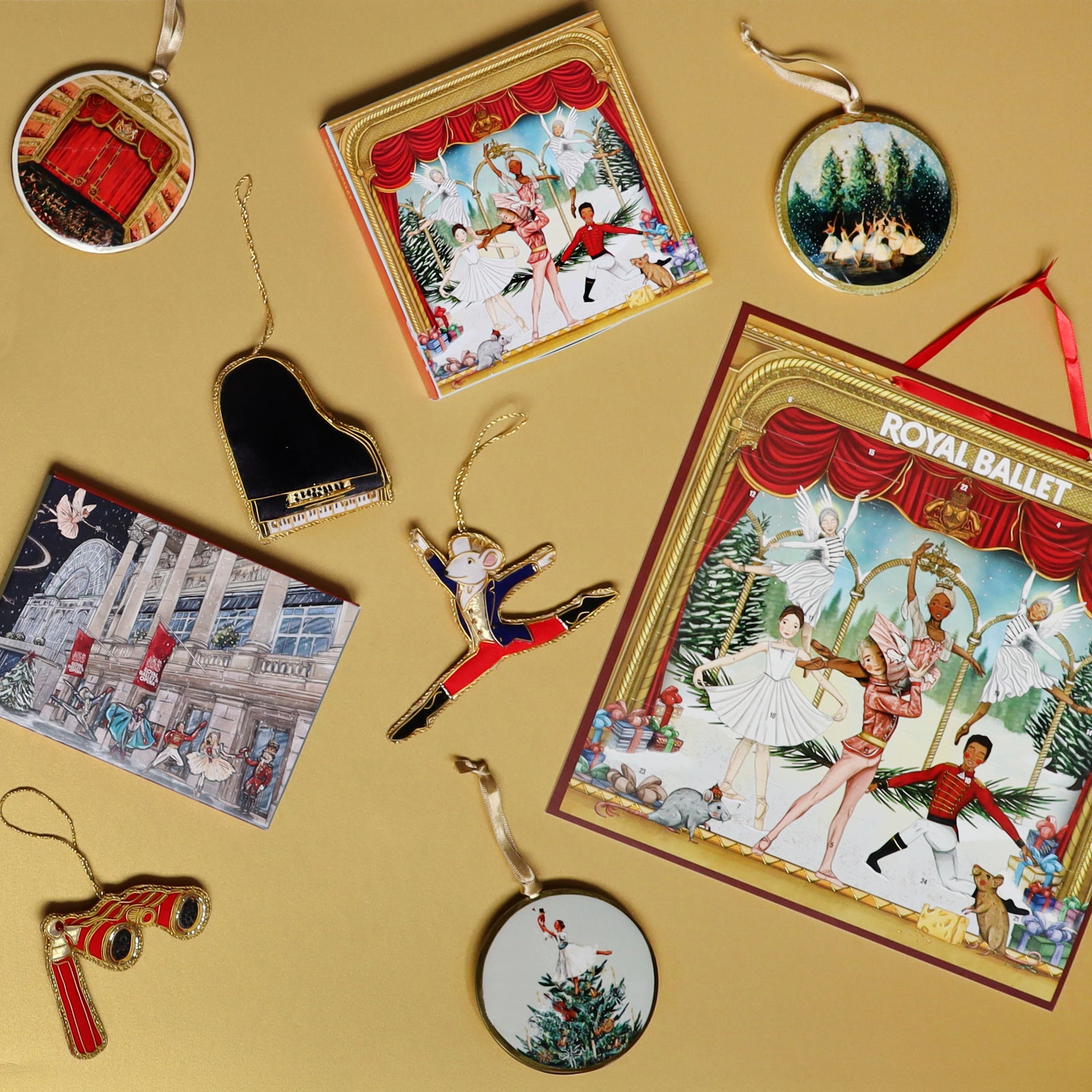 A collection of Royal Ballet Nutcracker-themed Christmas decorations, advent calendar and a matching cards arranged on a warm gold background. The set includes enamel tin decorations, a grand piano decoration, a leaping Mouse King, opera glasses, and an advent calendar featuring Nutcracker-style ballet dancers under red curtains.