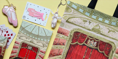 A tote bag with a detailed illustration the stage and curtains at the Royal Opera House, styled in a flat-lay alongside ballet shoes patch and decoration, a notebook, and playing cards on a yellow background.