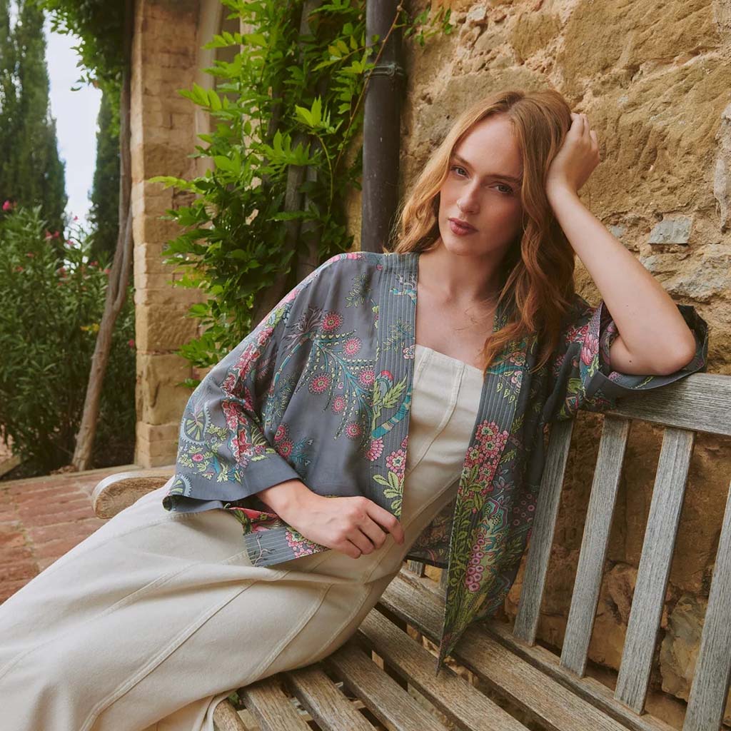 Woman in a floral kimono style lightweight jacket sitting on a wooden bench against a stone wall.