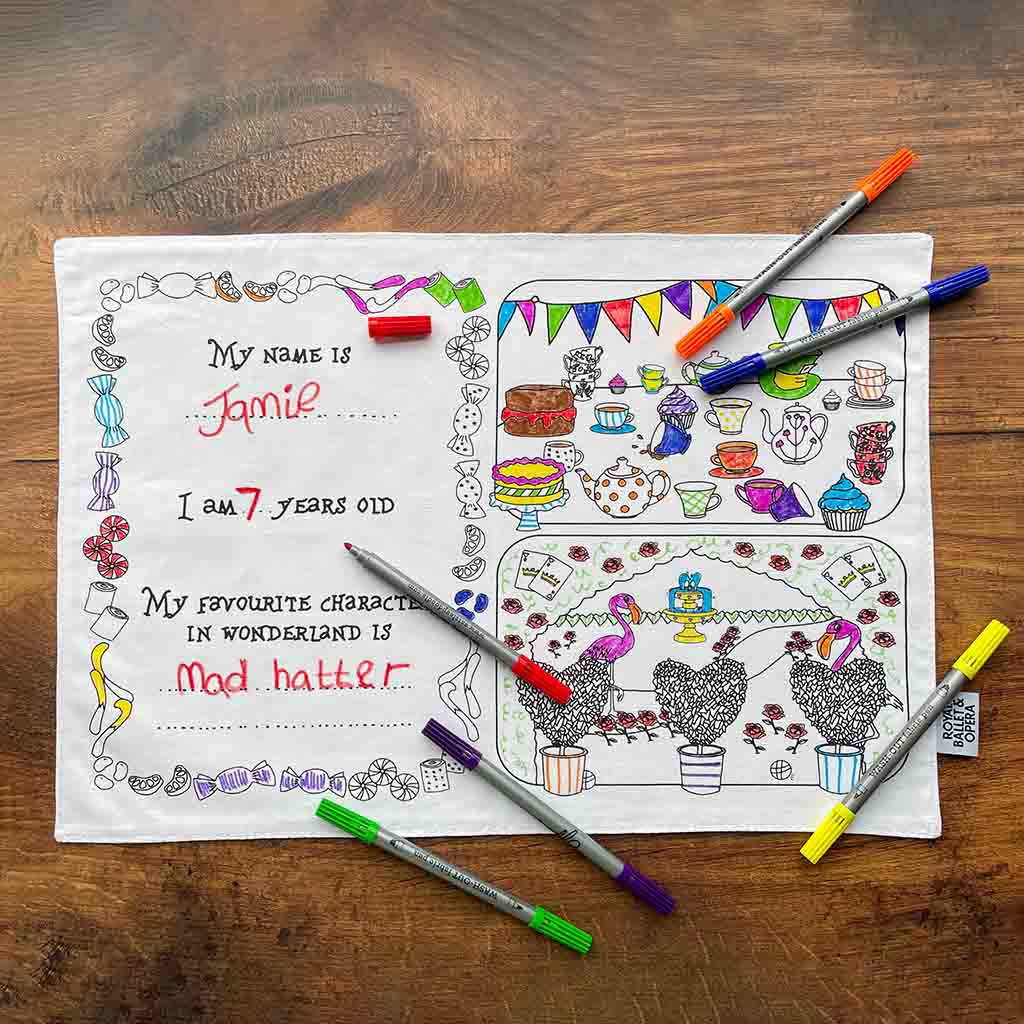 The back of a RBO Colour in Placemat against a wooden background with six different coloured pens scattered on top. The left side of the mat has text which reads &quot;My name is blank&quot;, &quot;I am blank years old&quot;, and &quot;My Favourite character from wonderland is blank&quot; with spaces to write answers. There is a border of sweets around this. Right side of the mat has two images, the top of a series of teacups and pots, as well as cakes and a top hat, and the bottom is of a garden with flamingos and a fountain. 