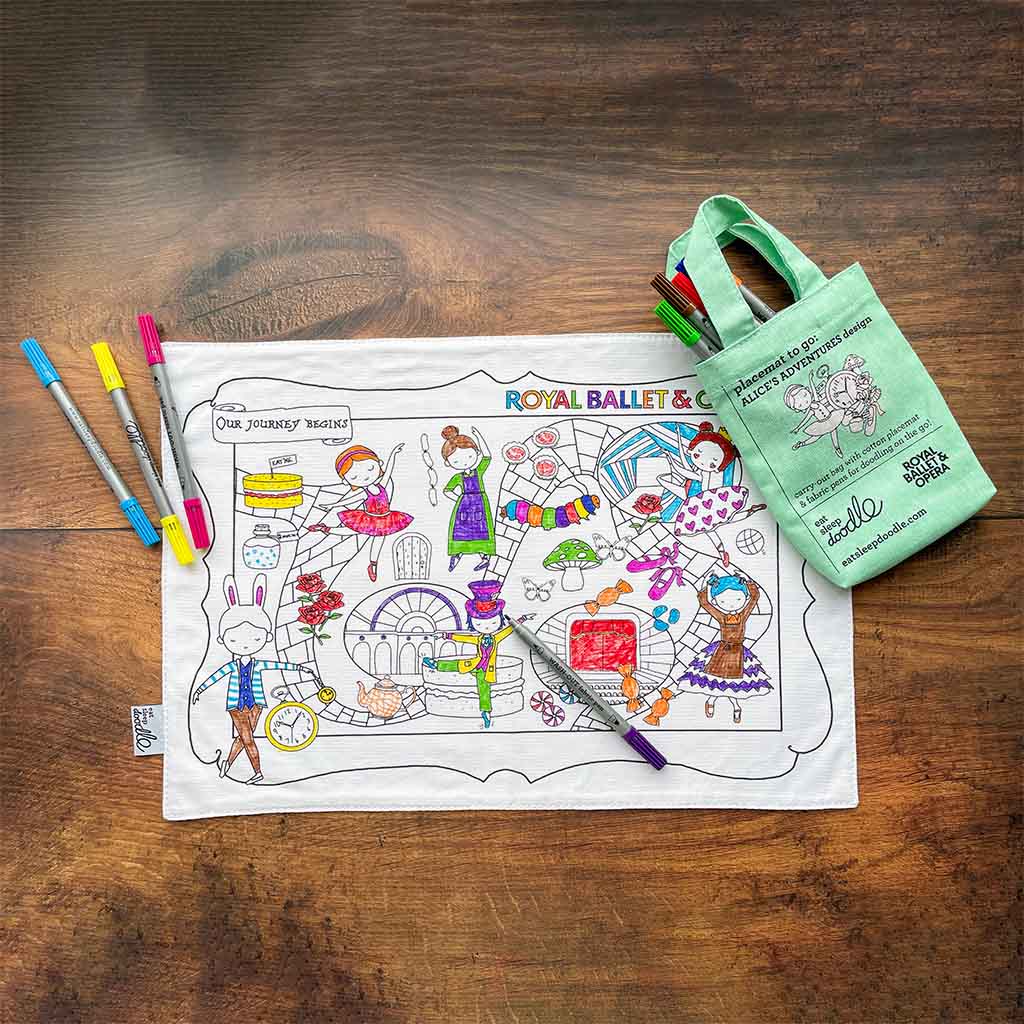 The front of a RBO Colour In Placemat against a wooden background with four coloured pens scattered on top and a green carrying bag with more pens protruding. 