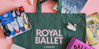 A dark green tote bag with 'ROYAL BALLET LONDON' printed in pink on the front, styled in a flat lay with a ballet shoes jigsaw, books, and a white swan hairclip on a pink background.