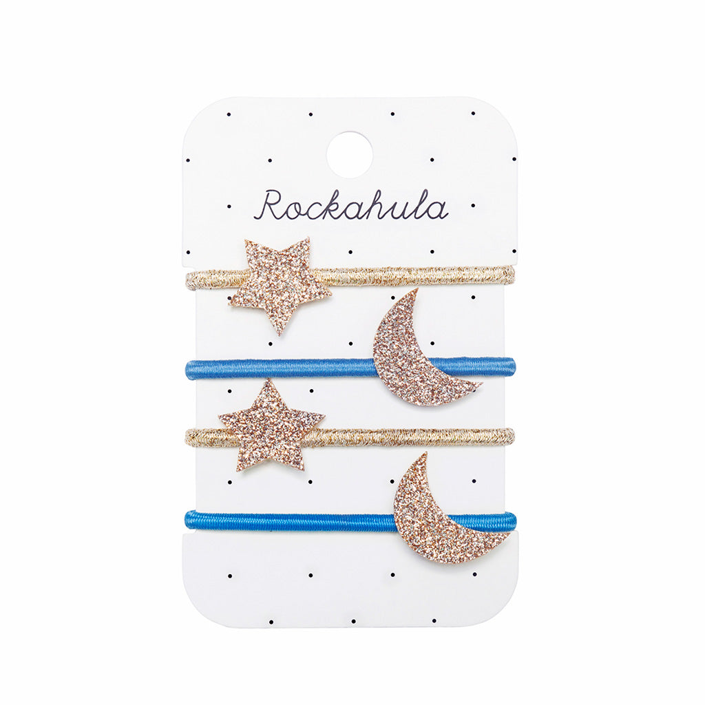 A set of Starry Skies Hair Ties on a white card with black dots against a white background. There are four elastic ties, two gold and two blue. The gold ties have a glittery star attached and the blue ties have a glittery crescent moon attached. 