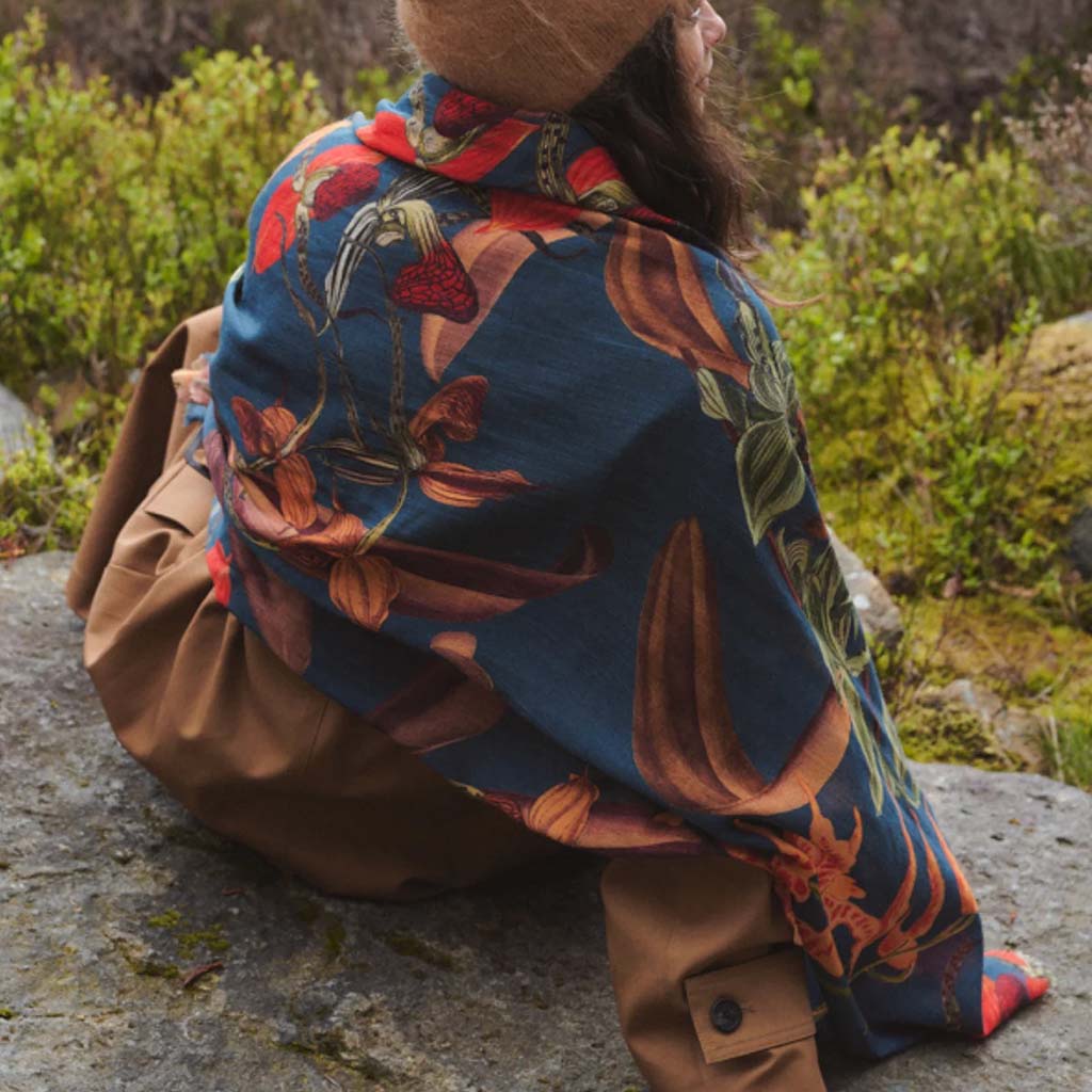 back view of a model sitting outside in nature with a brown hat and coat, with teal blue and orange wool scarf 
