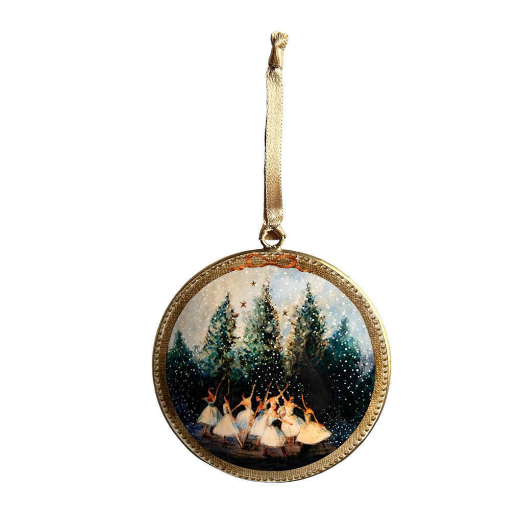 A Snowflakes Tin Decoration designed by Elena Deshmukh against a white background. The decoration is round and has a bronze ribbon attached by a small metal ring. The design features a gold frame around the outside of the decoration with a bow painted on at the top. Within the frame is a snowy scene where 8 female figures in white dresses are dancing in front of pine trees. We can see 3 stars in the sky behind the trees. 