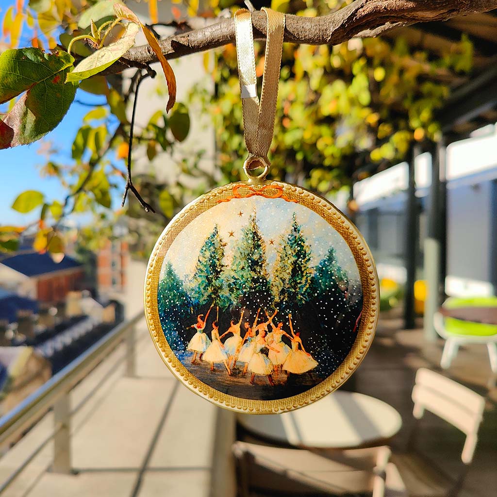 A Snowflakes Tin Decoration hanging from a leafy branch. The background is out of focus but we can see a terrace balcony with tables and chairs laid out and there is more greenery in the background. We can see over the balcony on the left of the image that this is several floors up as we can see the tops of buildings. It is a sunny day. 