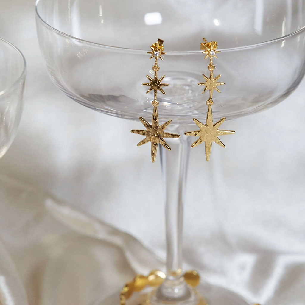 A pair of gold starburst dangle earrings hanging from a clear champagne glass on a soft white fabric background.