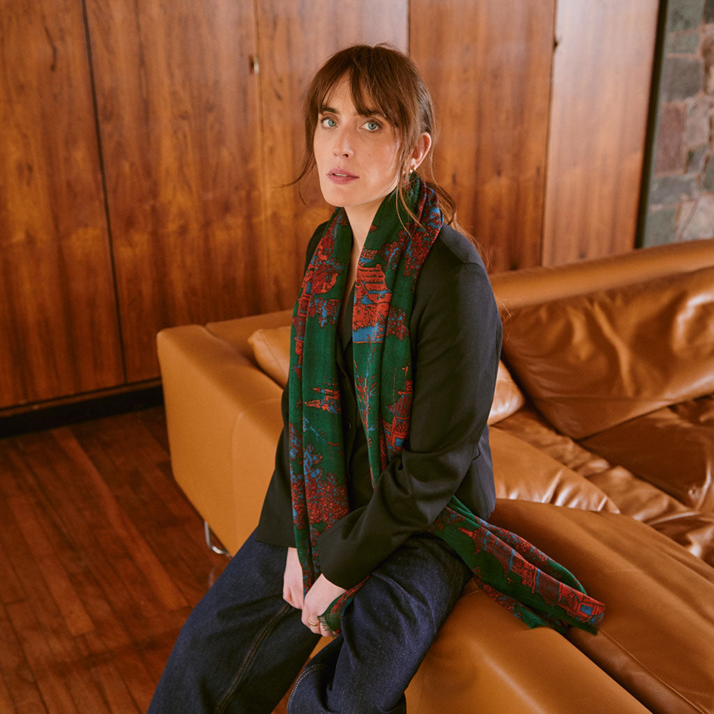 Woman sitting on a brown leather couch wearing a green and red Pagoda Forest scarf in a wooden-paneled room.