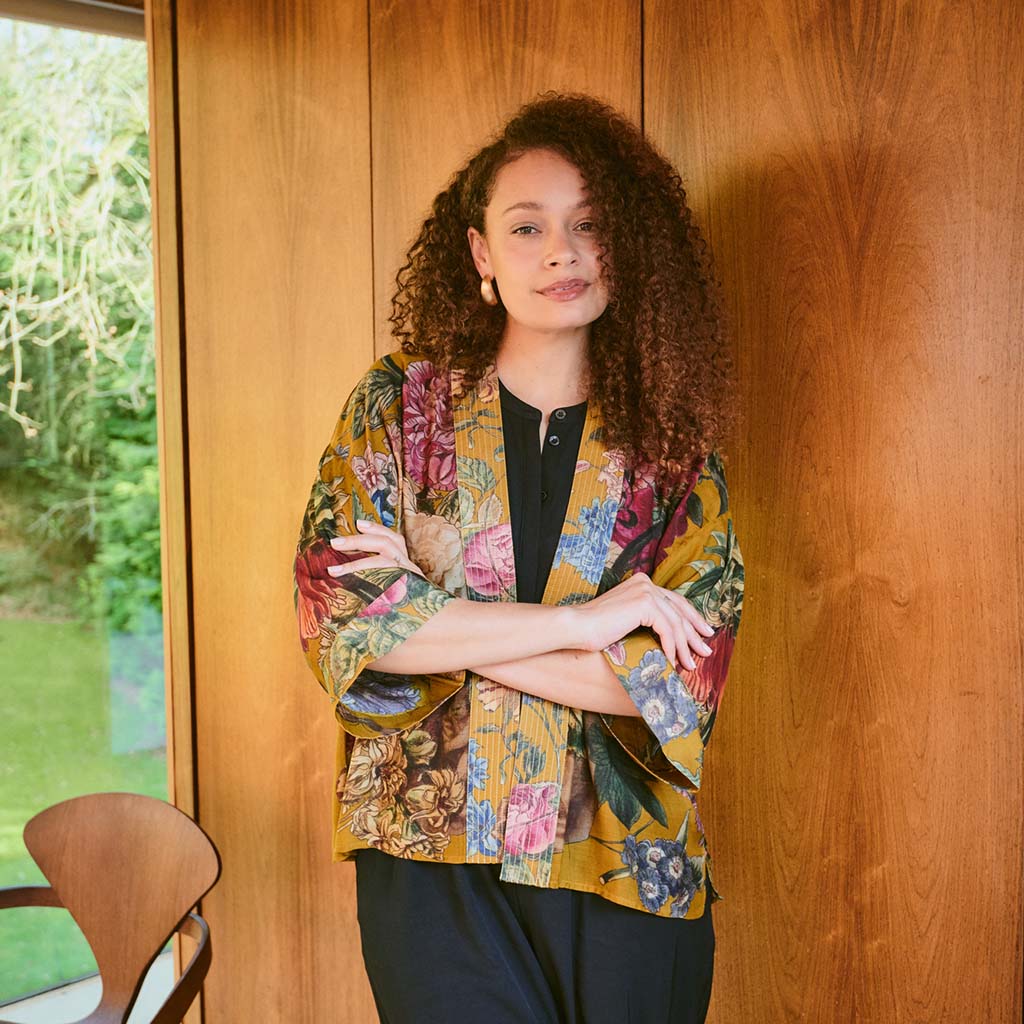 Woman wearing a colorful floral  kimono style jacket standing against a wooden wall.