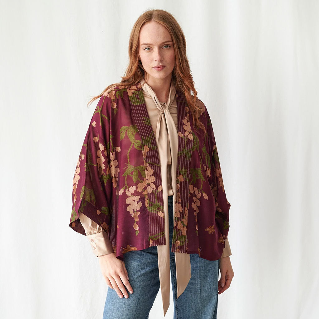 A woman stands against a white curtain backdrop wearing a burgundy  plum kimono-style jacket with a beige and green floral wisteria print, layered over a cream blouse and paired with blue jeans.