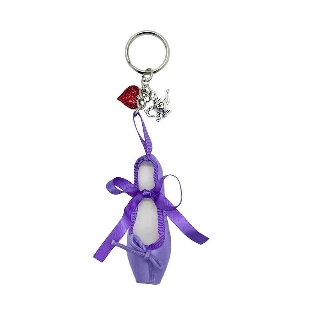 A Limited Edition Alice Ballet Shoe Keying against a white background. It is comprised of a purple keyring in the shape of a ballet shoe. The keyring is hanging vertically, suspended by a short metal chain with a larger ring at the top for attaching to keys. Attached to the chain there are two metal charms, one in the shape of a teapot with a heart etched on, and one is a red heart. There are two purple bows, a larger one about two thirds of the way up the shoe, and a smaller one at the bottom. 