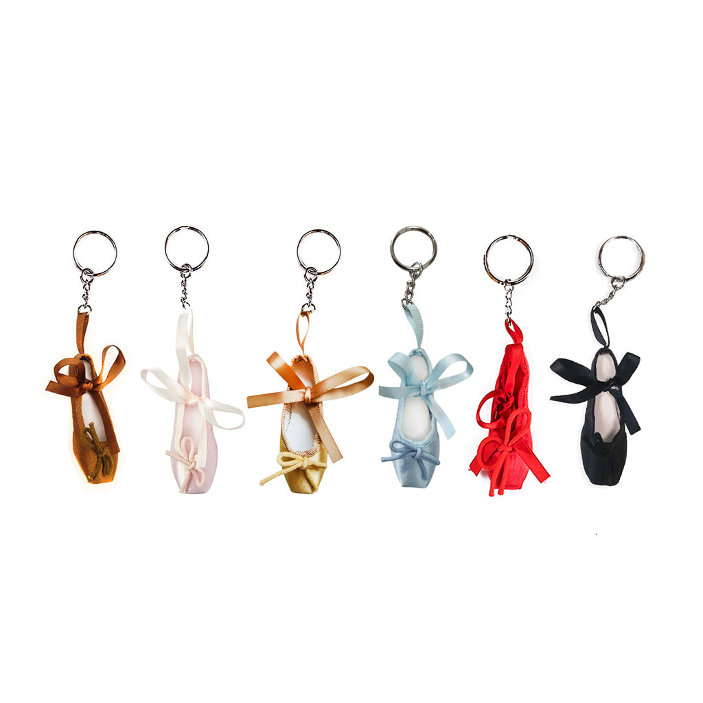 A selection of six ballet shoe keyrings in various colours. From left to right we can see bronze, pink, pale gold, blue, red, and black. They are against a white background and are hanging vertically with their chains above them. 