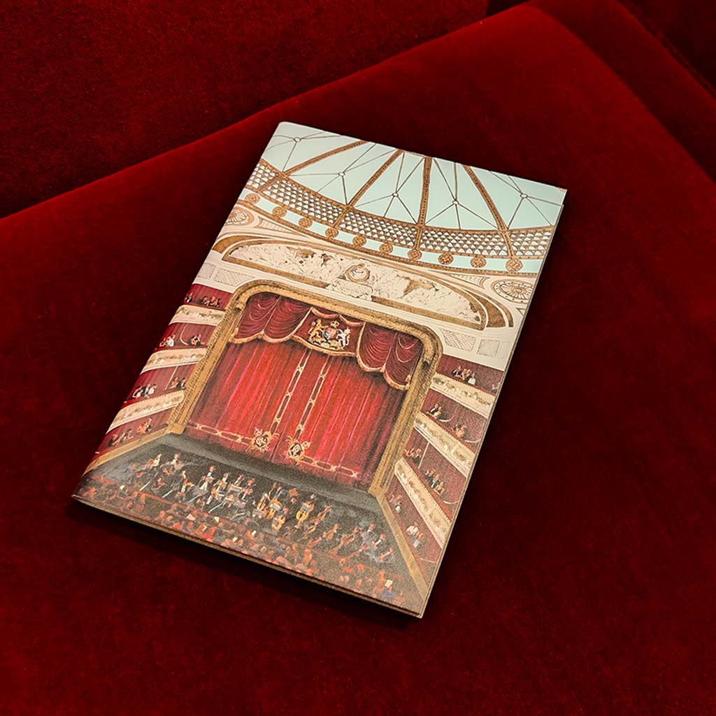 A leather covered notebook featuring an illustration of the auditorium at the Royal Opera House  with red curtains and an audience, placed on a plush red velvet surface.
