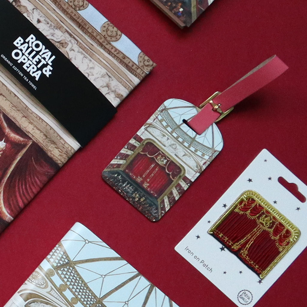 A flat-lay product photo of a Royal Ballet &amp; Opera Auditorium themed products including a luggage tag, tea towel and iron on patch. The set includes an illustrated red velvet opera house curtain, all on a deep red background