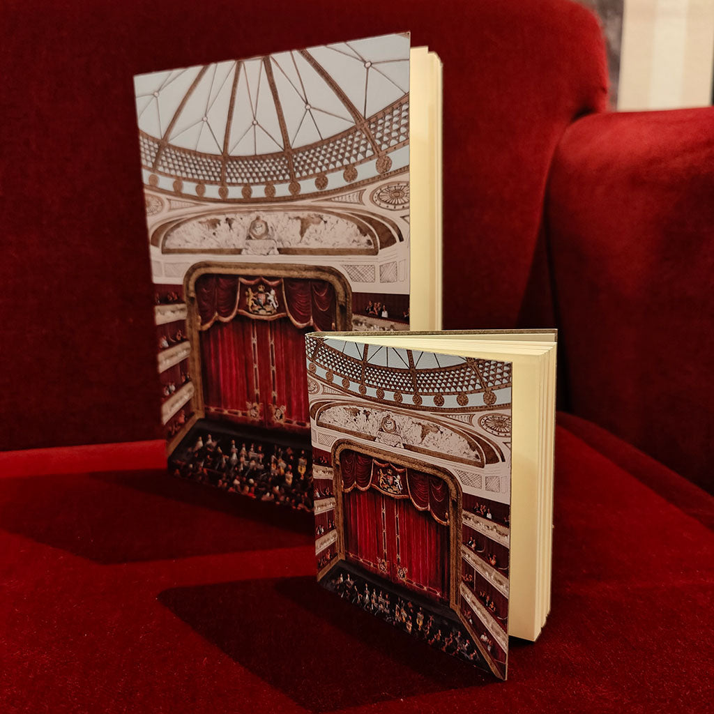 Two leather notebooks A5 and mini versions with illustrated covers depicting the auditorium at the Royal Opera House with the stage and balcony, standing on a red velvet surface in front of a matching red velvet background.