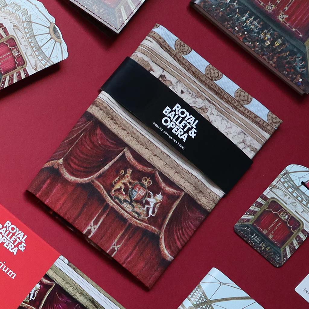 A Royal Ballet &amp; Opera organic cotton tea towel lies on a red surface, surrounded by other auditorium products. The tea towel features a black band with white text reading 'ROYAL BALLET &amp; OPERA' and an illustrated red stage curtain and gold decorative motif.