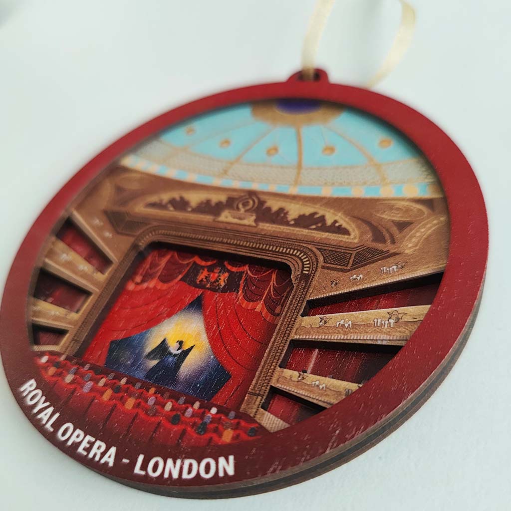 A round wooden hanging decoration featuring an illustrated view of the Royal Opera House in London, with red curtains, tiered seating, and a hanging loop at the top.