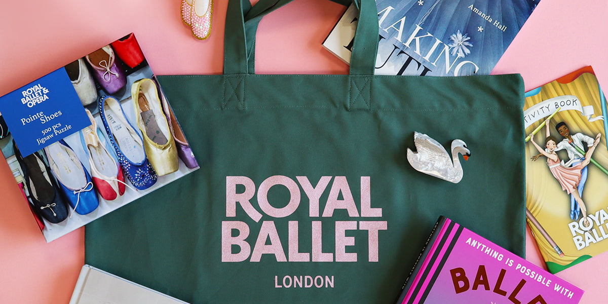 A dark green Royal Ballet London tote bag laid flat on a pink background, surrounded by ballet-related items including a 'Pointe Shoes' jigsaw, a swanclaw clip, and other ballet-themed books.