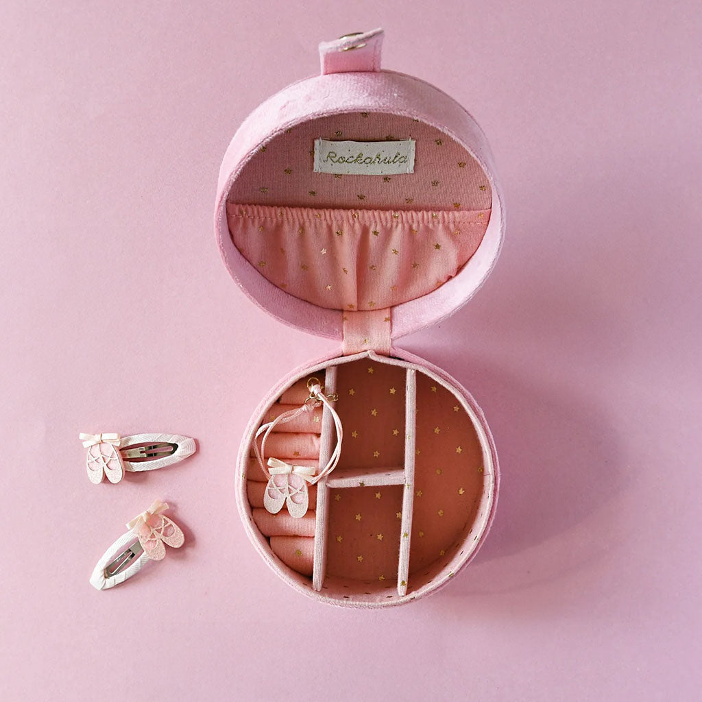 An open Mini Ballet Jewellery Box  on a pink background. The box is shop open to reveal a pink and gold star fabric lined interior. The bottom of the box is divided into 4 compartments, in one of which is a small ballet shoe charm attached to a thread bracelet. Next to the box are 2 pink ballet shoe hairclips. 