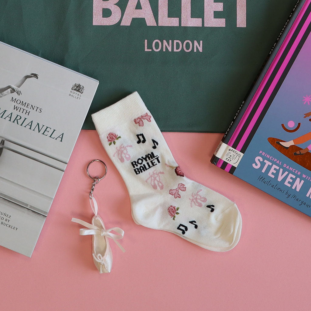 Pair of socks with ballet-themed designs on a pink surface with books and a green bag in the background.