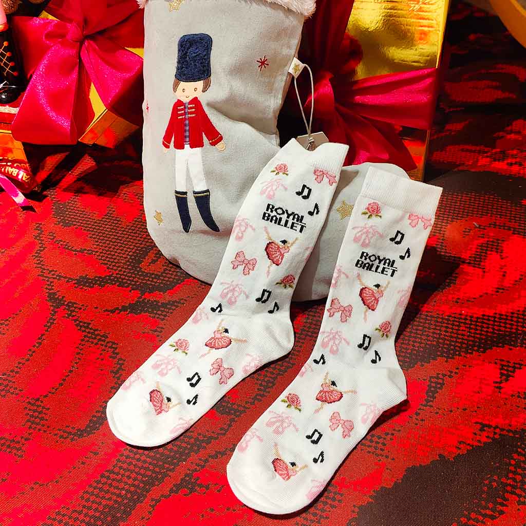White socks with ballet-themed designs on a red and black patterned surface, with a decorative bag in the background.