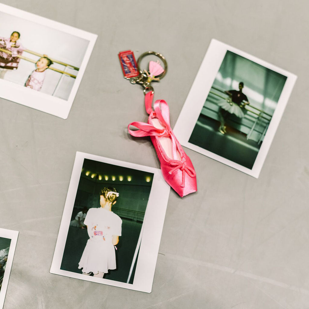A pink satin Barbie Ballet Shoe keyring with two charms styled on a gray dance studio floor alongside several Polaroid photos.