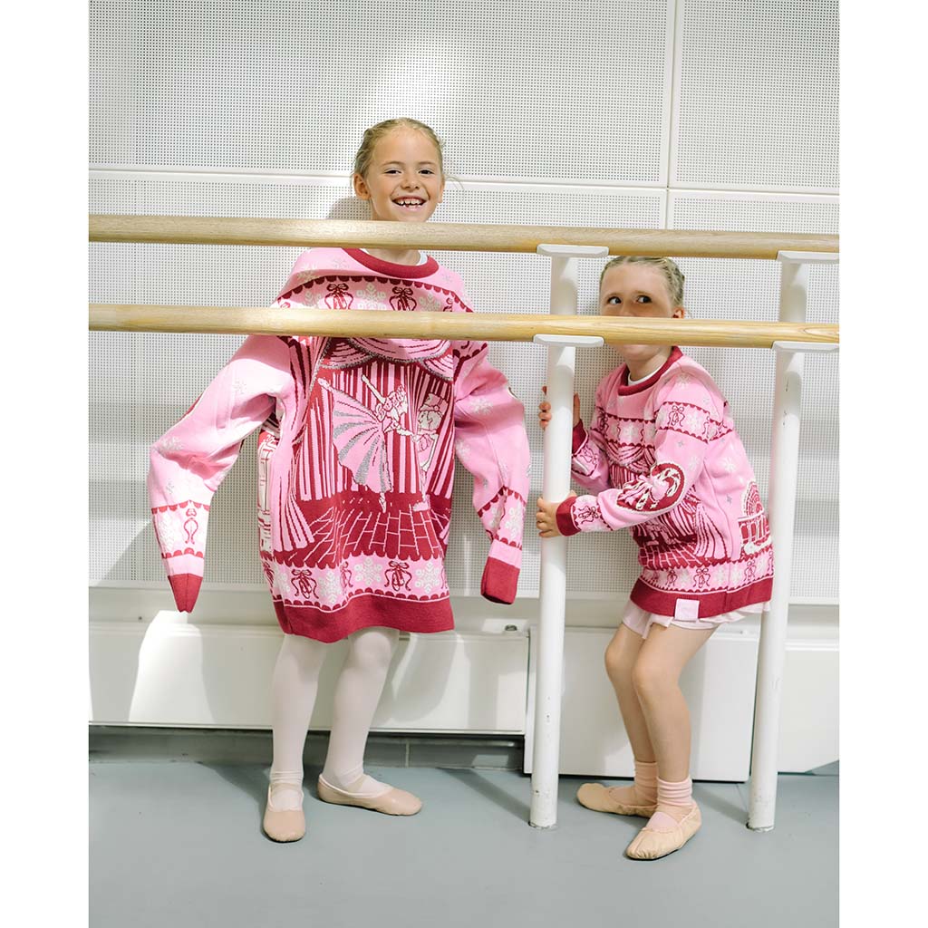 Two children in a ballet studio playing behind a ballet barre wearing pink Barbie Royal Ballet Christmas Jumpers one is wearing an adult version which is too big
