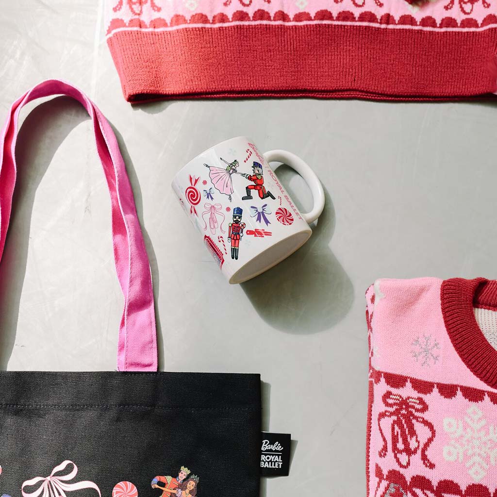 A styled flat-lay featuring a white ceramic mug with a colourful Barbie Royal Ballet theme print, a black tote bag with pink straps and a visible Barbie Royal Ballet tag, and partial views of pink christmas jumper on a light gray dance studio floor.