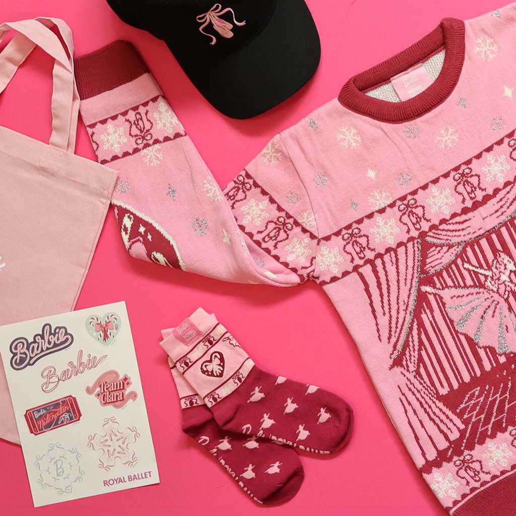 A styled flat-lay of a pink Barbie Royal Ballet Christmas Jumper with a ballet theme, matching pink and burgundy patterned socks, a black baseball cap with ballet shoe embroidery, a light pink tote bag, and a Barbie-themed sticker card on a bright pink background.