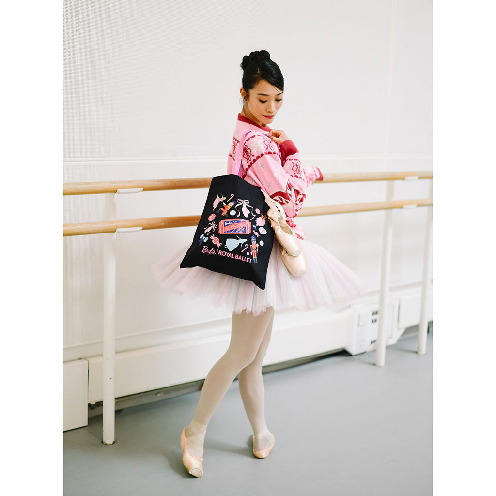 A ballerina in a pink barbie Christmas Jumper and white tutu holds a black tote bag featuring a colourful ballet illustration and the text 'Barbie Royal Ballet'.
