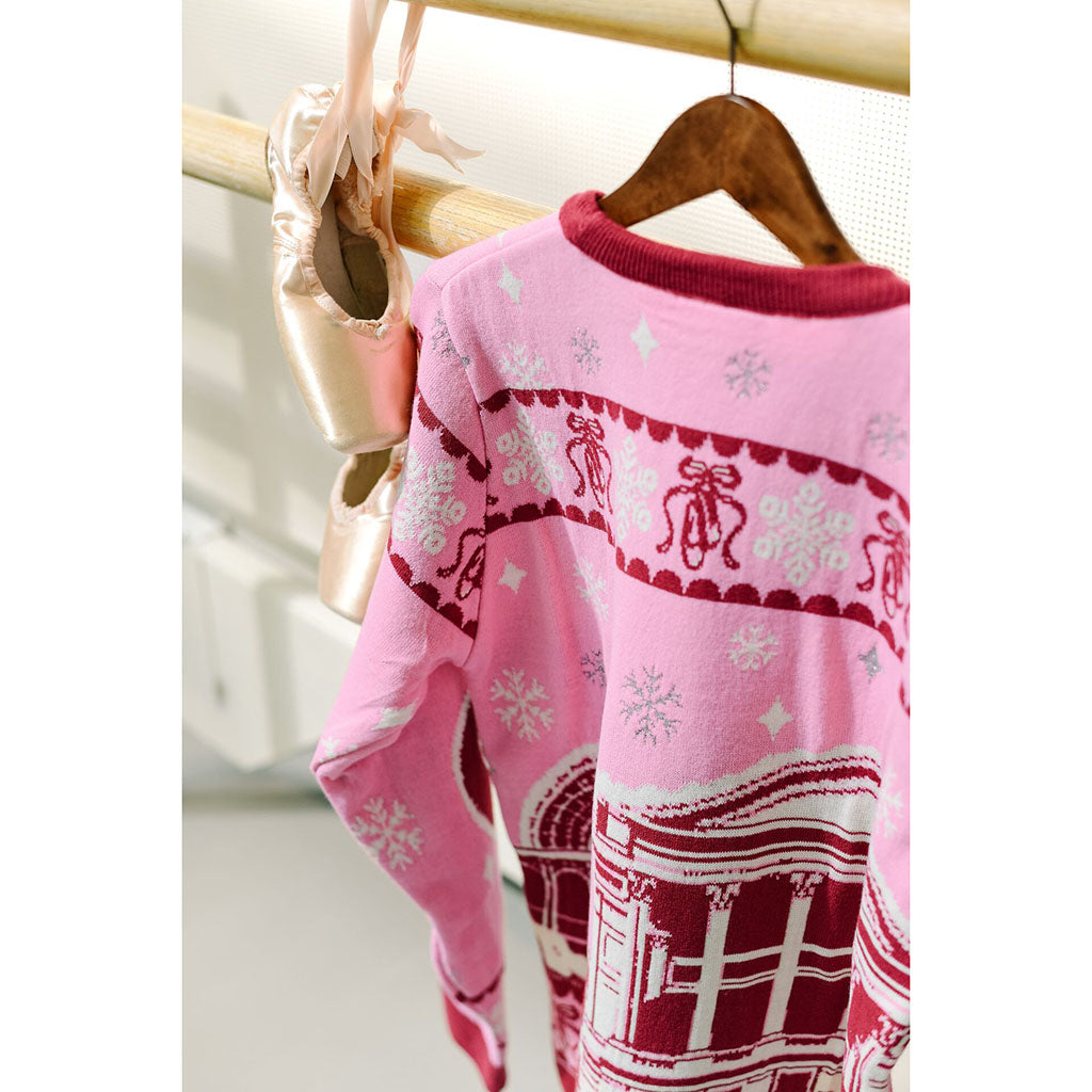 A view of the back of a pink long-sleeve Barbie Christmas Jumper with a holiday pattern featuring snowflakes, bows, and a red-and-white Royal Opera House graphic, hanging on a wooden hanger next to ballet pointe shoes