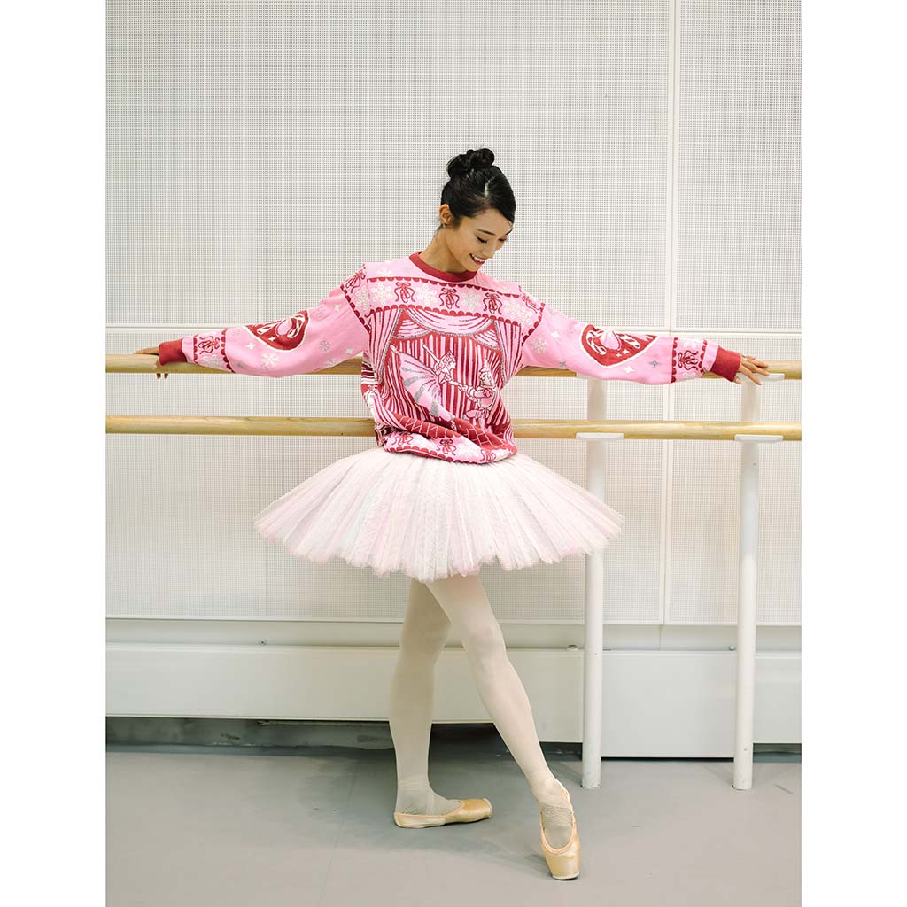 Principal dancer Fumi Kaneko posing in a ballet studio wearing a pink and red patterned long-sleeve christmas Barbie Jumper, a white tutu, white tights, and pointe shoes, leaning against a wooden barre