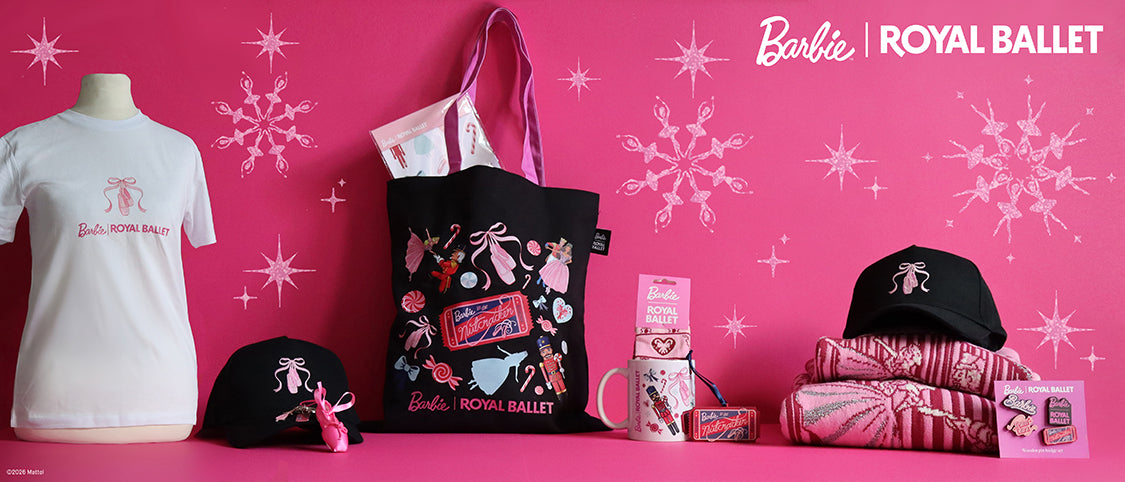 A Barbie Royal Ballet merchandise display on a bright pink background with snowflake graphics. The display includes a white T-shirt on a mannequin, a black tote bag with pink handles and colourful ballet-themed print, a black cap, folded pink Barbie Christmas Jumper, a mug, and small accessories.