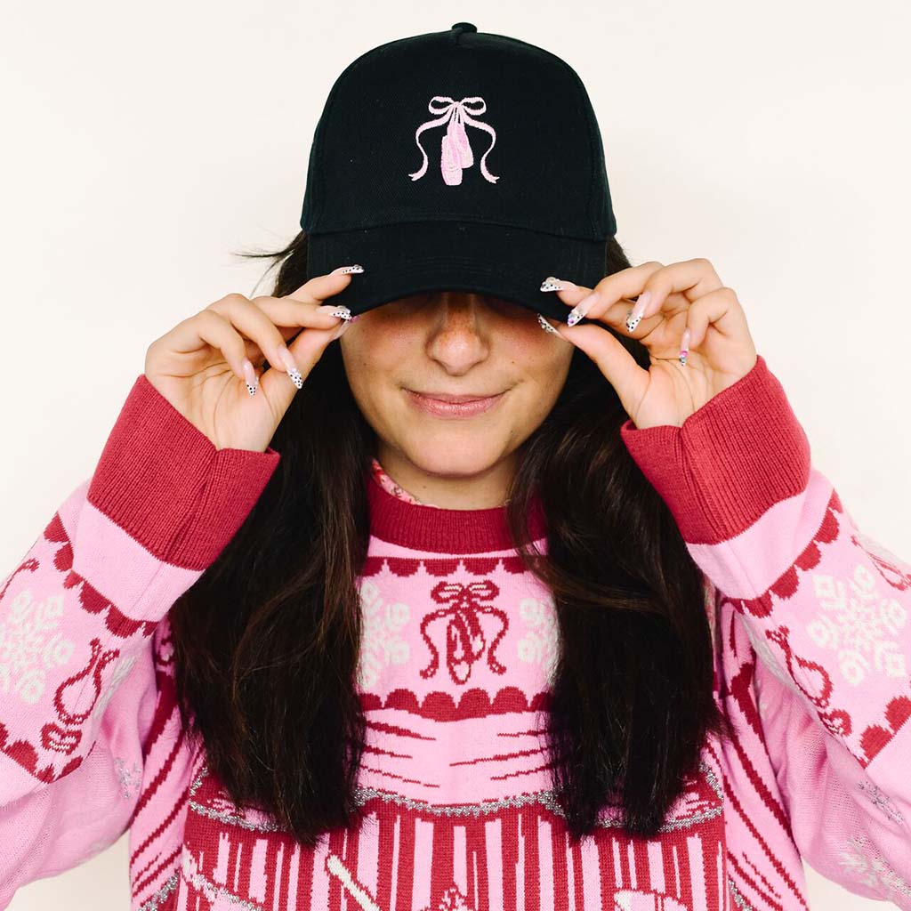 A woman wearing a black baseball cap with a pink pointe shoe and ribbon embroidered on the front, holding the brim over her eyes. She is wearing a pink Barbie Christmas Jumper with red patterned against a plain off-white background.