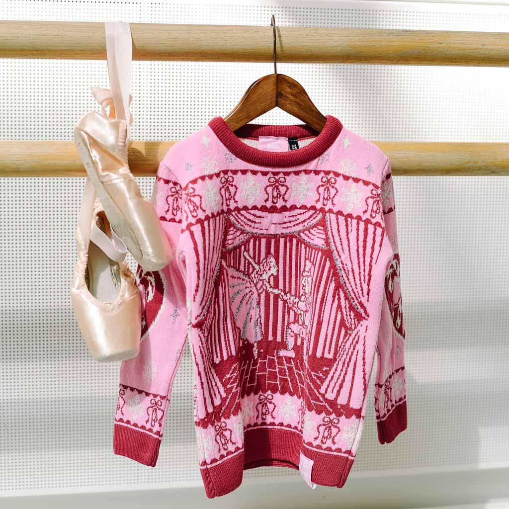 A pink Barbie Royal Ballet Christmas Jumper with a dark pink graphic of a Clara and the Nutcracker on a stage, featuring patterned cuffs and hem hanging on a ballet barre with pointe shoes in a studio