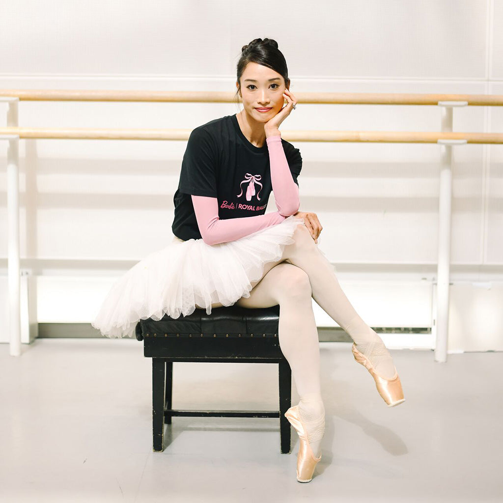 Principal Dancer Fumi Kaneko in a ballet studio sits on a black  piano stall, wearing a black graphic t-shirt over a long sleeved top and a white tutu, paired with beige tights and pink pointe shoes. The t-shirt features pink ballet shoes and text Barbie Royal Ballet.