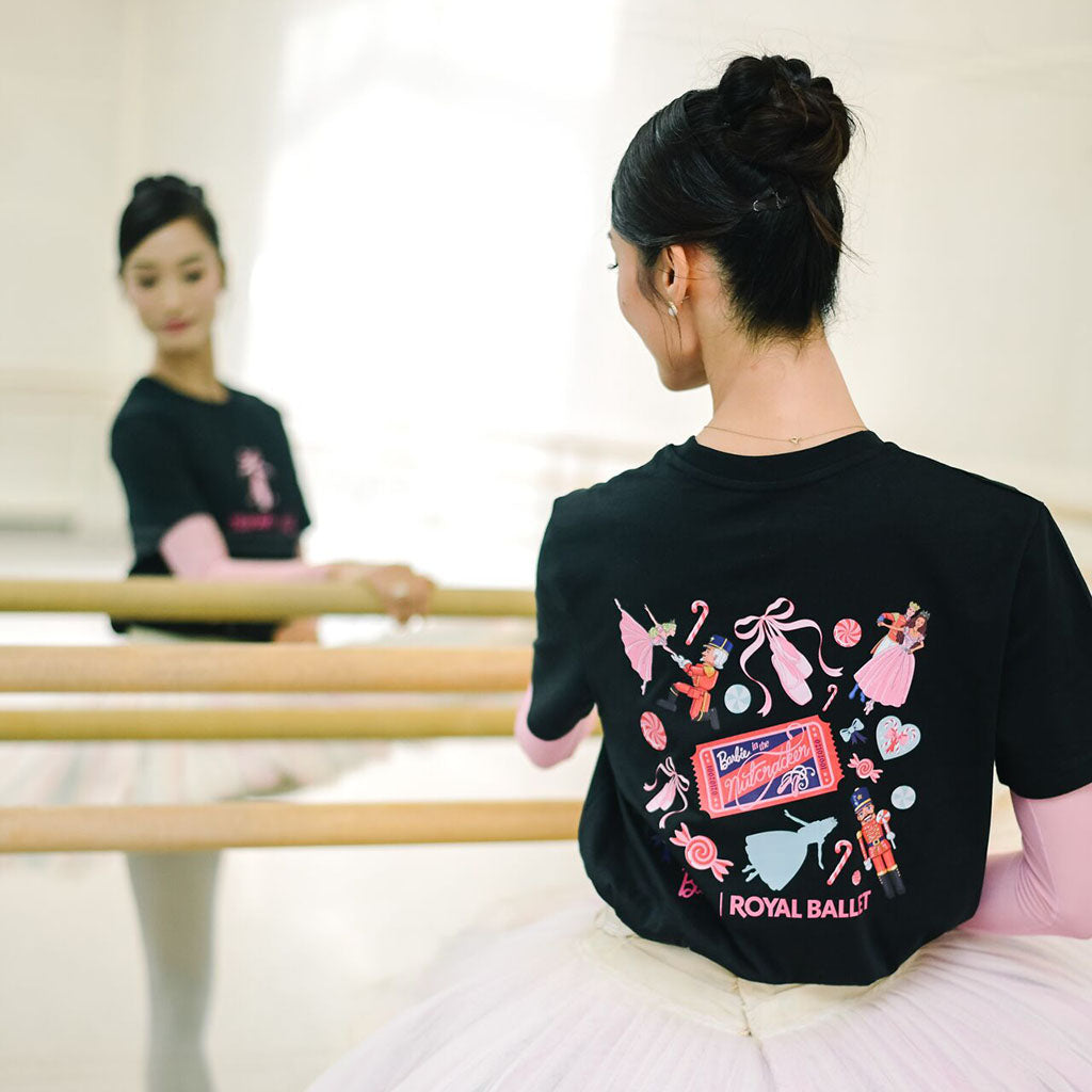 Principal Dancer Fumi Kaneko in a ballet tutu seen from behind wearing a black short-sleeve t-shirt with a colourful back graphic that reads 'Barbie ROYAL BALLET'. She is in a bright dance studio with wooden barres, with her reflection visible in the mirror.