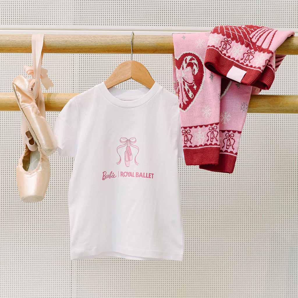 A children's white short-sleeve T-shirt with a pink ballet shoe and ribbon graphic and 'Barbie ROYAL BALLET' text, hanging on a ballet barre. A pink patterned Barbie Christmas Jumper and a pair of nude pointe shoes hang beside it against a light textured wall.