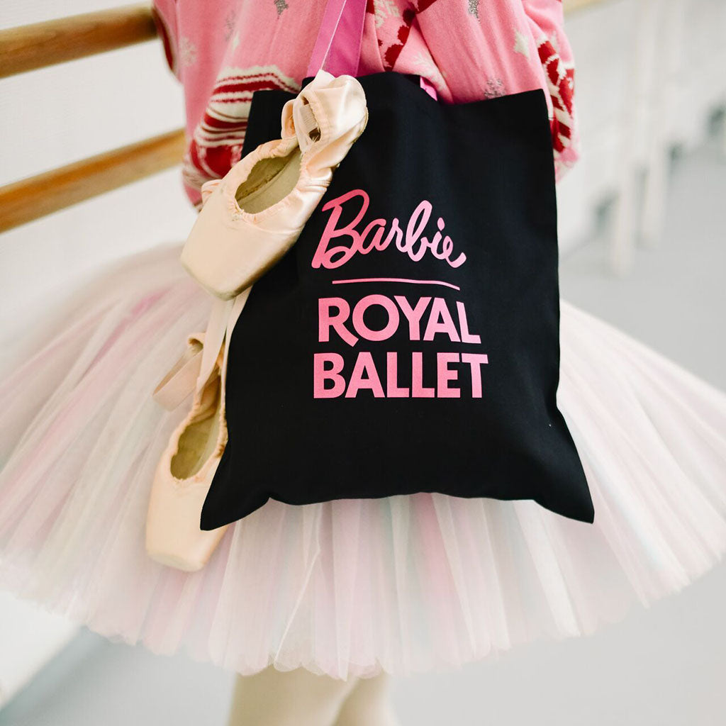 the reverse of the Barbie Tote black with 'Barbie ROYAL BALLET' in pink lettering, held by principal dancer Fumi wearing a pink ballerina outfit and tutu with pink pointe shoes.