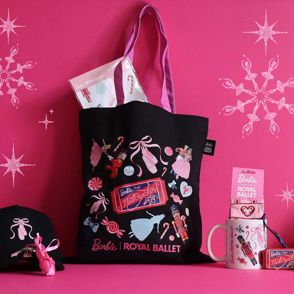 A black tote bag with pink handles and Barbie Royal Ballet graphics, displayed on a pink surface against a pink background with snowflake decals. The bag is styled with a matching black cap, a pink ballet shoe keyring, a ticket decoration, and a white mug and socks with Barbie Royal Ballet artwork.