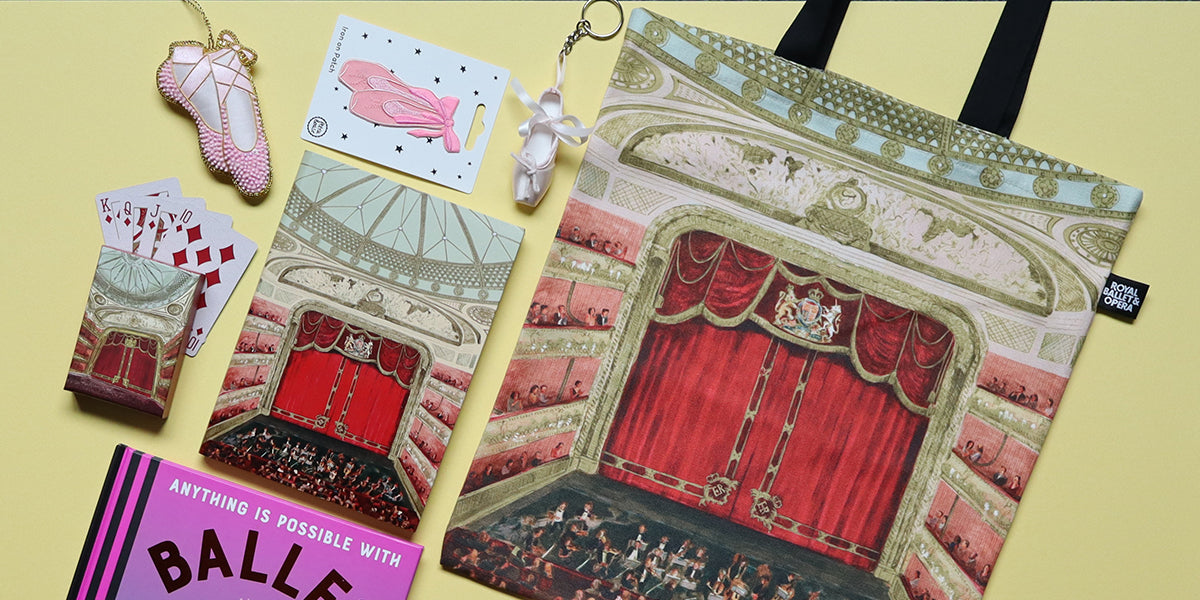 A tote bag featuring an illustrated red theatre stage and auditorium, styled in a flat lay with ballet-themed accessories on a pale yellow background.