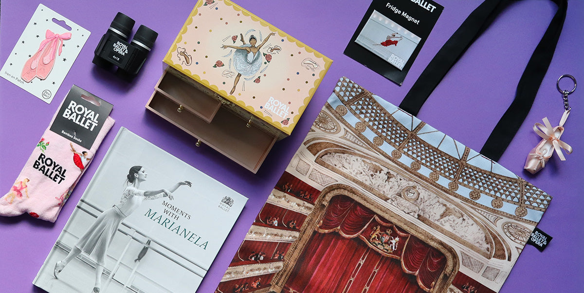 A curated flat-lay of ballet-themed merchandise on a solid purple background, including a printed tote bag, a ballet jewellery box, a pair of Royal Ballet socks, a ballet book, and other accessories.