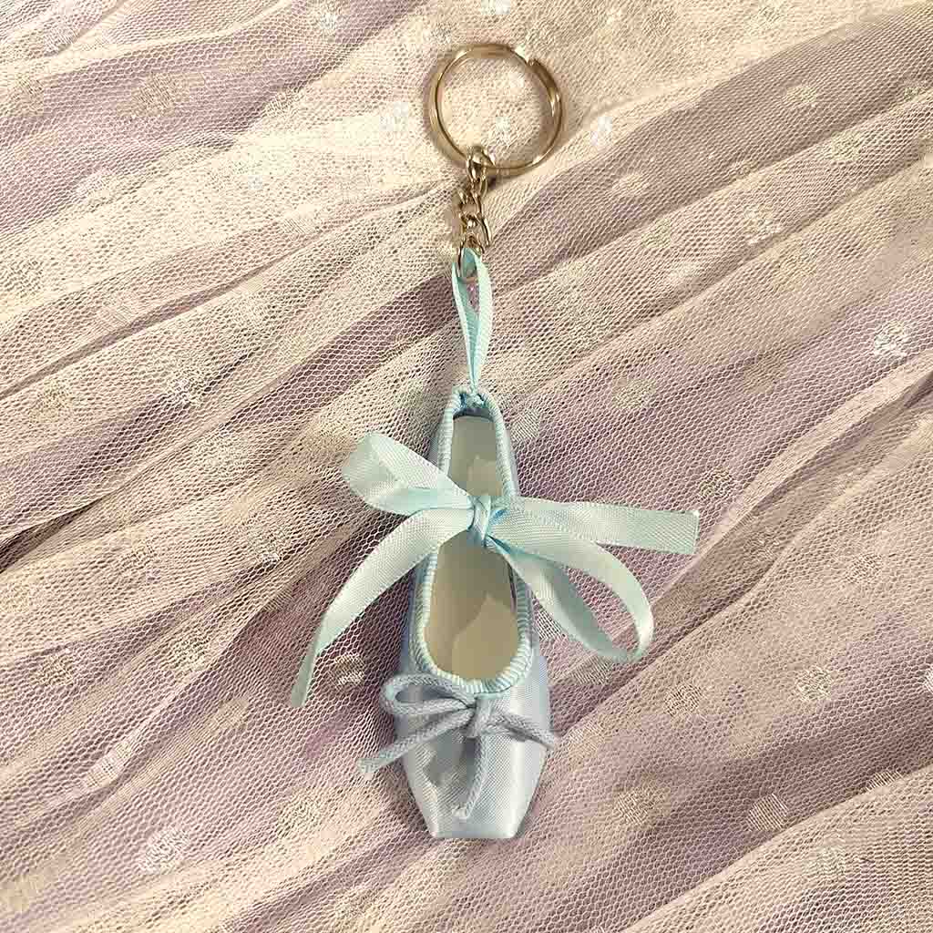 A blue keyring in the shape of a ballet shoe against a white background. The keyring is hanging vertically, suspended by a short metal chain with a larger ring at the top for attaching to keys. There are two blue bows, a larger one about two thirds of the way up the shoe, and a smaller one at the bottom. 