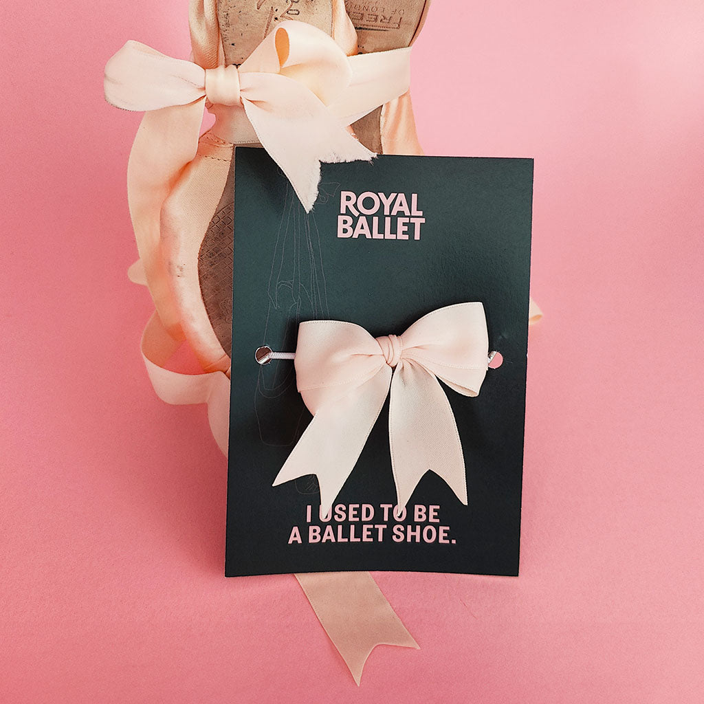 A pale blush pink satin bow hair clip displayed on a black card that reads 'ROYAL BALLET I USED TO BE A BALLET SHOE.' against a solid pink background.