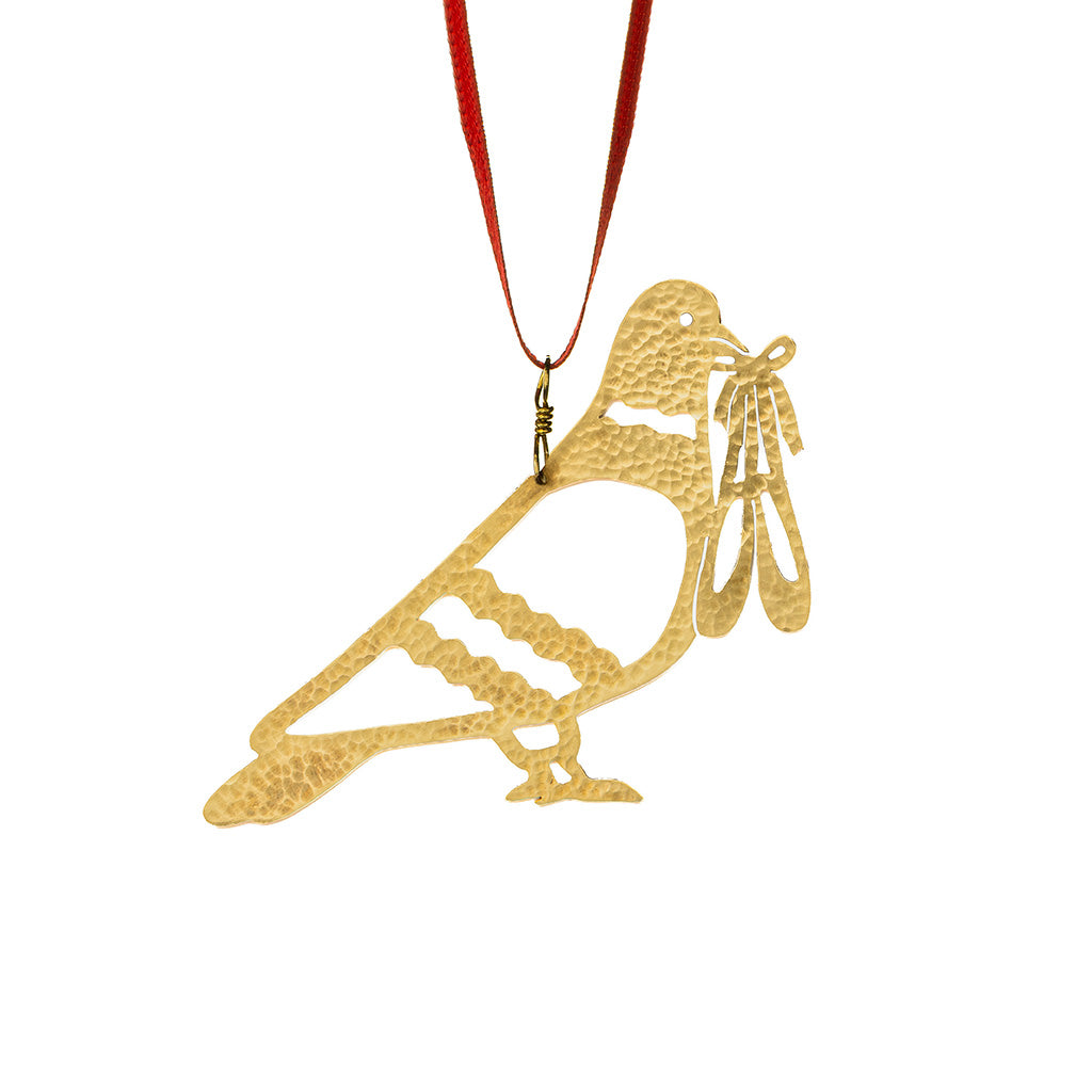 brass decoration featuring a silhouette of a pigeon holding ballet pointe shoes in its beak hanging from a red ribbon on a white background