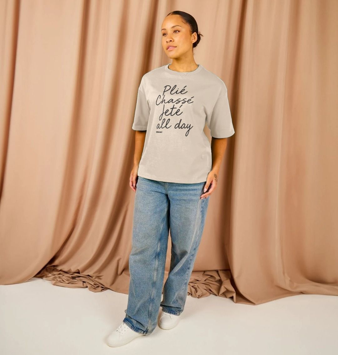 Person wearing a oat t-shirt with text Plié, Chassé, Jeté all day and blue jeans against a beige curtain background