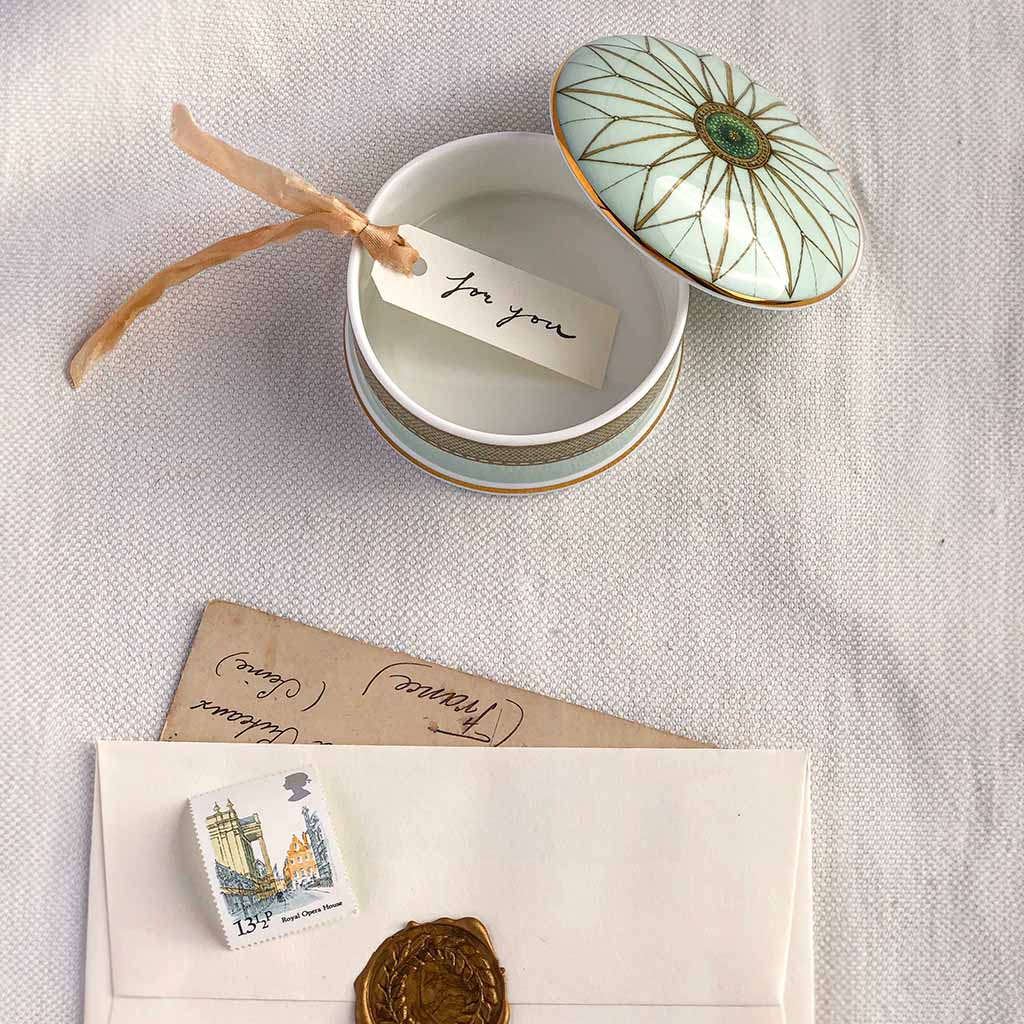 An Auditorium Ceiling Trinket Box open and on display on a white cloth background. Inside is a white label with &#39;for you&#39; hand written in ink and a bronze ribbon attached. At the bottom of the page is a brown paper letter underneath a white envelope closed with a gold wax seal. There is a stamp on top of the envelope showing a street with the text &quot;13 1/2 p Royal Opera House&quot;. 