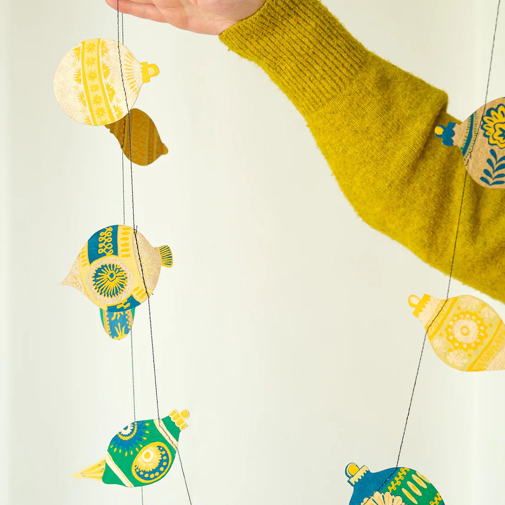 Colourful bauble-shaped paper and string garland hanging from a hand wearing a mustard yellow sweater.