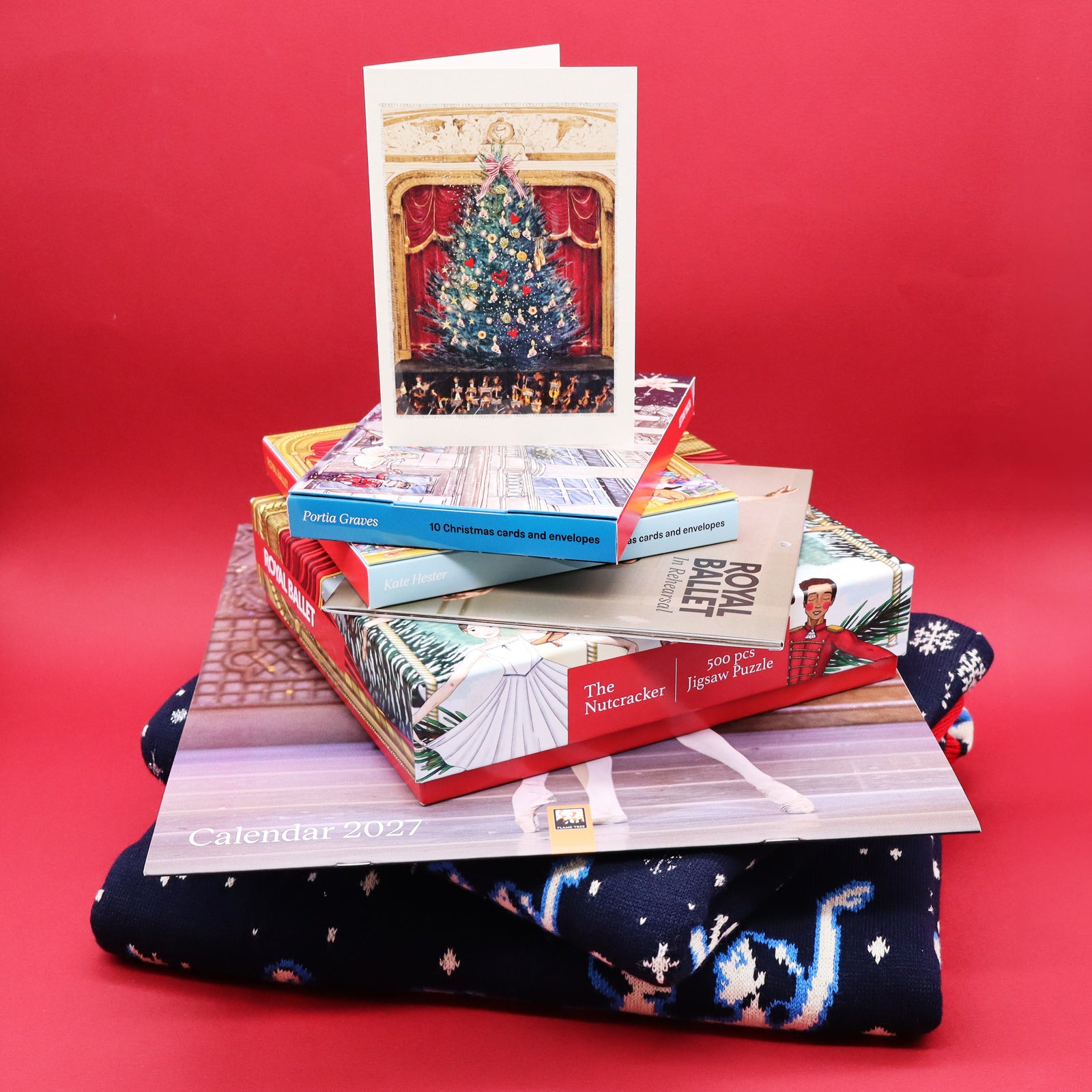 A stack of Christmas-themed paper goods and gift items, including a greeting card with a decorated Christmas tree, greetings card packs, a calendar, a Nutcracker jigsaw box, arranged on top of a dark navy patterned Christmas Jumper against a solid red background.